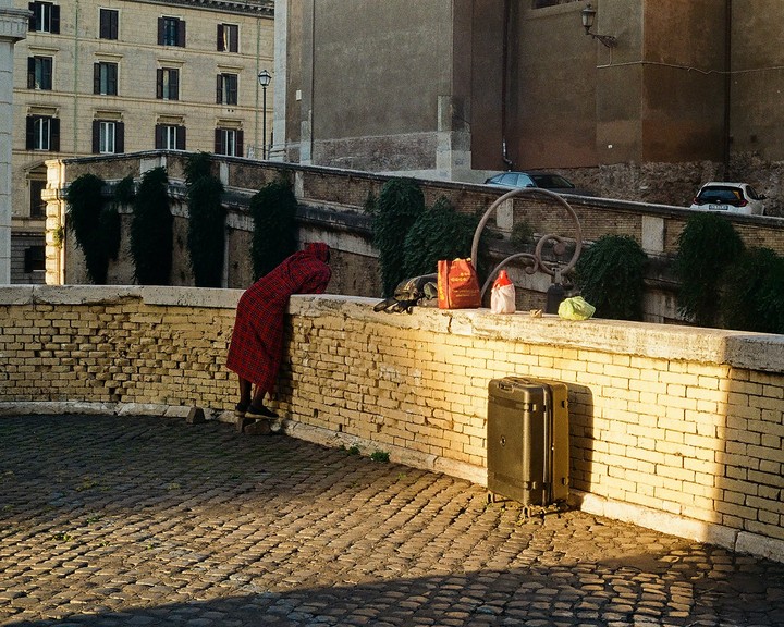 A person in a red robe leans over a brick wall next to a suitcase on a cobblestone street.