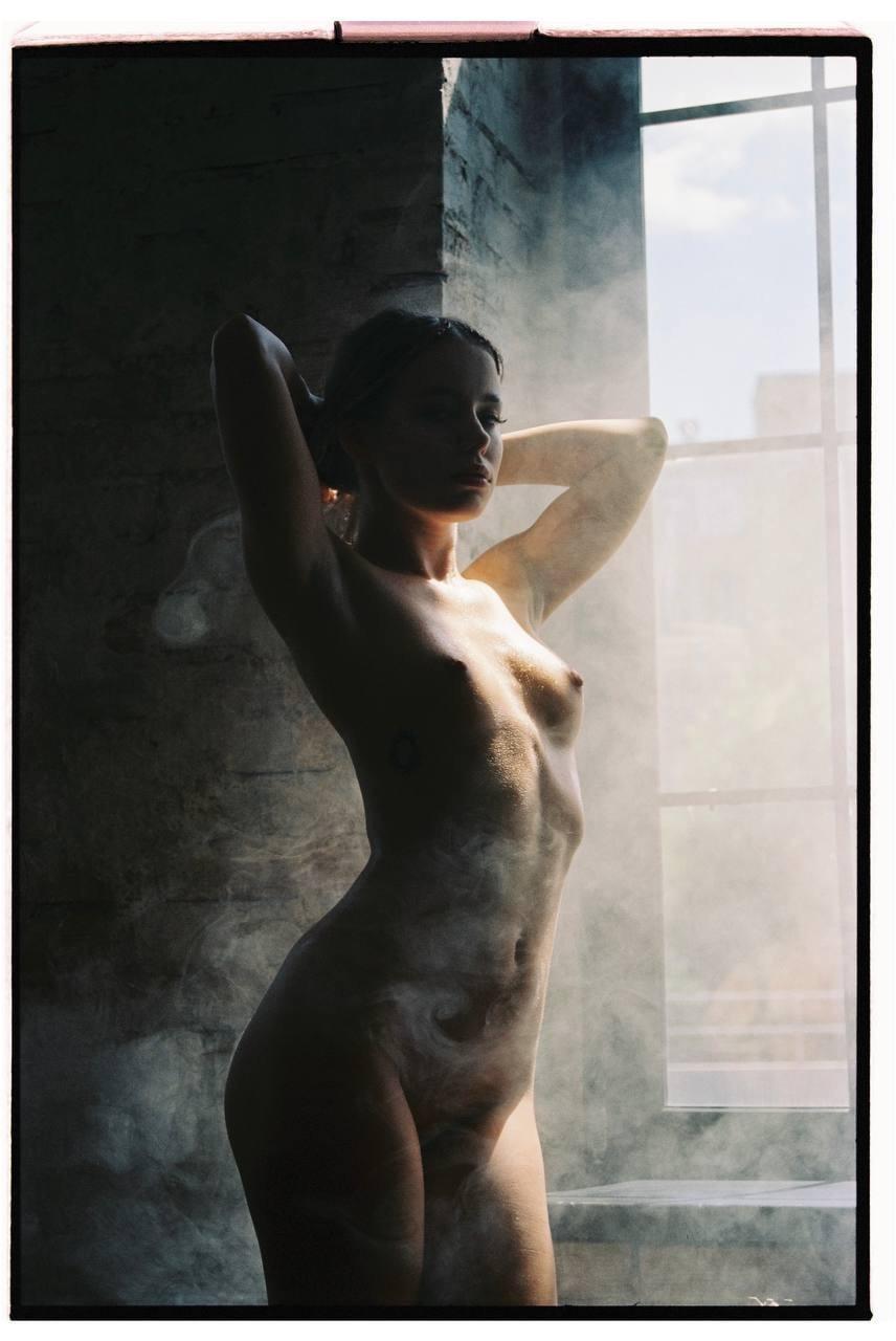 A woman stands nude with her arms behind her head, partially obscured by smoke, next to a window.