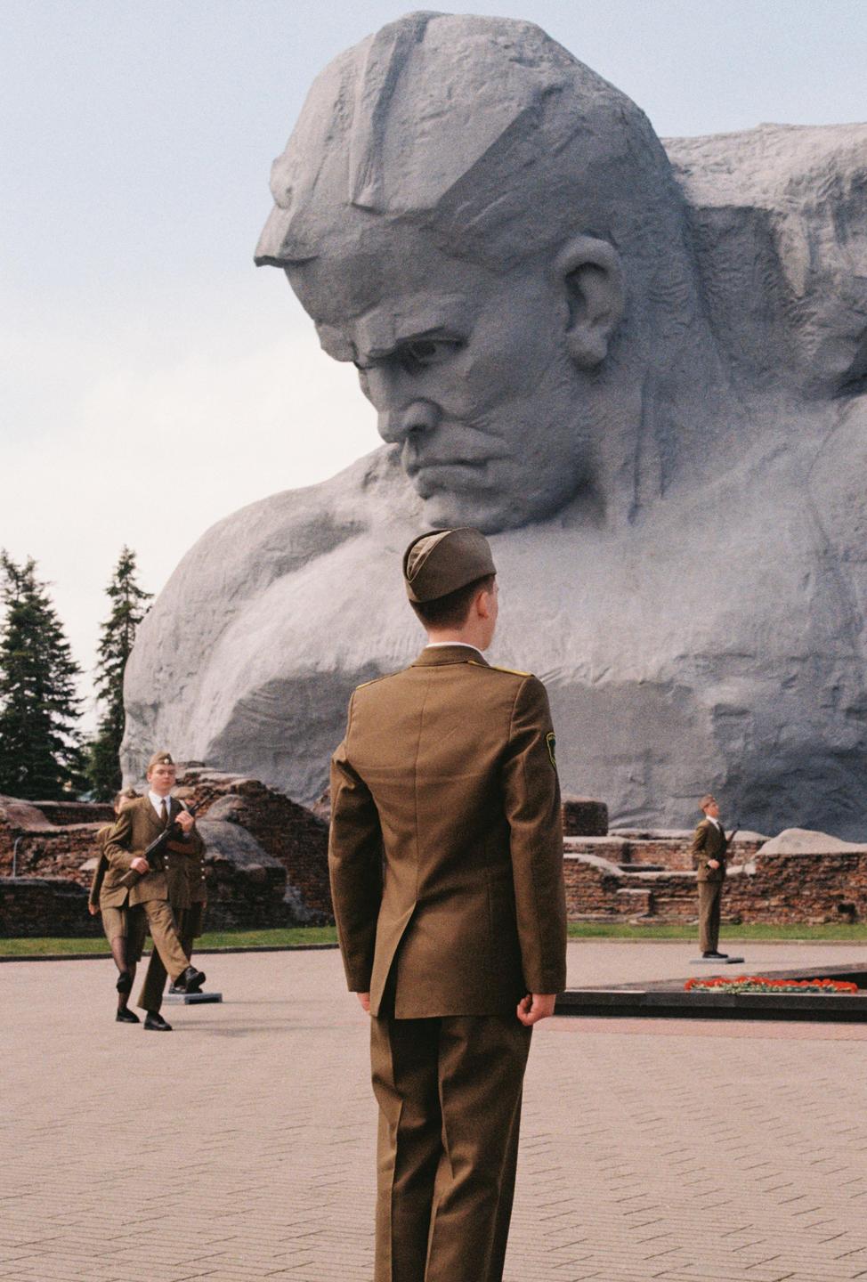 A man in uniform stands with his back to the camera, looking at a large stone sculpture of a man's head, with other men in uniform visible in the background.
