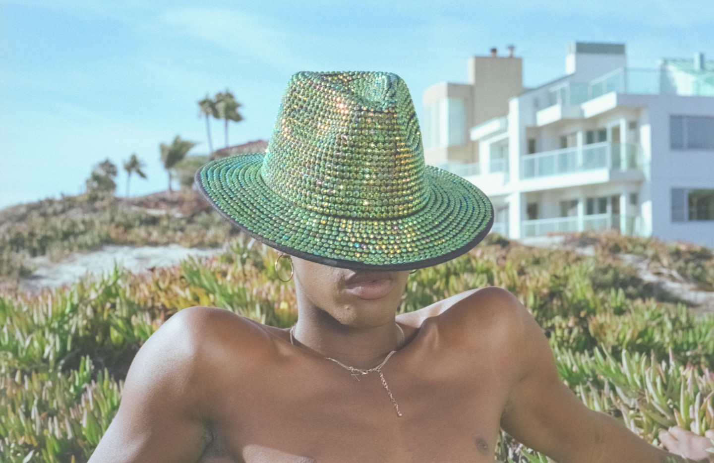 A person wearing a sparkly green hat that covers their face is pictured outdoors with green foliage and buildings in the background.