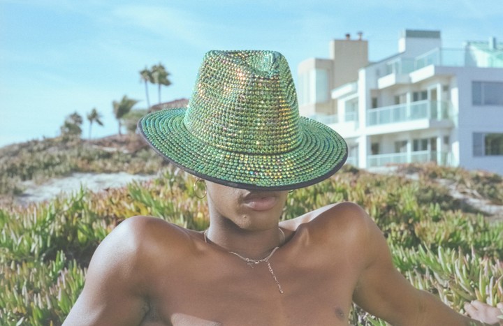 A person wearing a sparkly green hat that covers their face is pictured outdoors with green foliage and buildings in the background.
