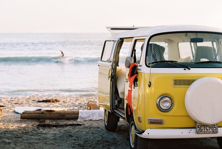 A yellow and white van with an open door is parked on a sandy beach with a surfer in the ocean.