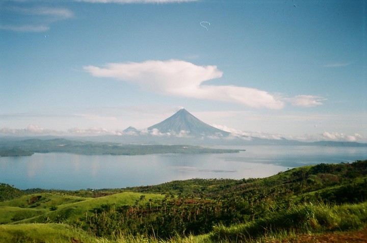A large, conical mountain rises from a body of water, surrounded by green hills and a blue sky with scattered clouds.