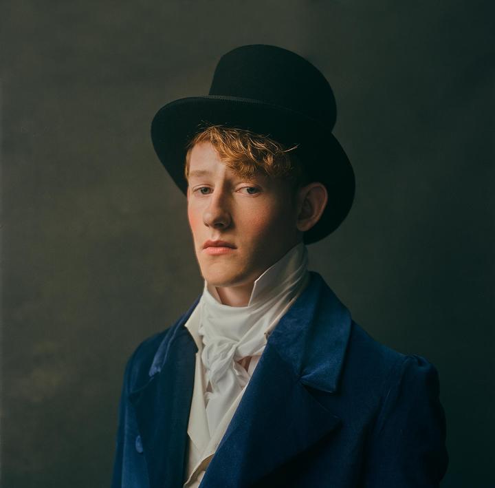 A man with red hair wears a top hat, a white cravat, and a blue velvet coat.