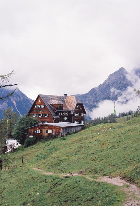 A large wooden building with green shutters sits on a grassy hill with mountains and clouds in the background.