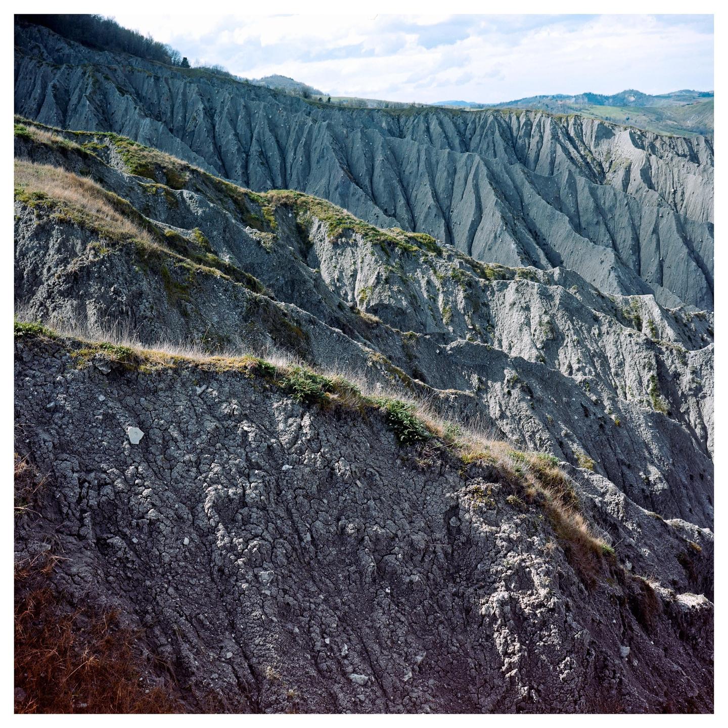 A landscape of badlands with steep, eroded hills and sparse vegetation under a cloudy sky.