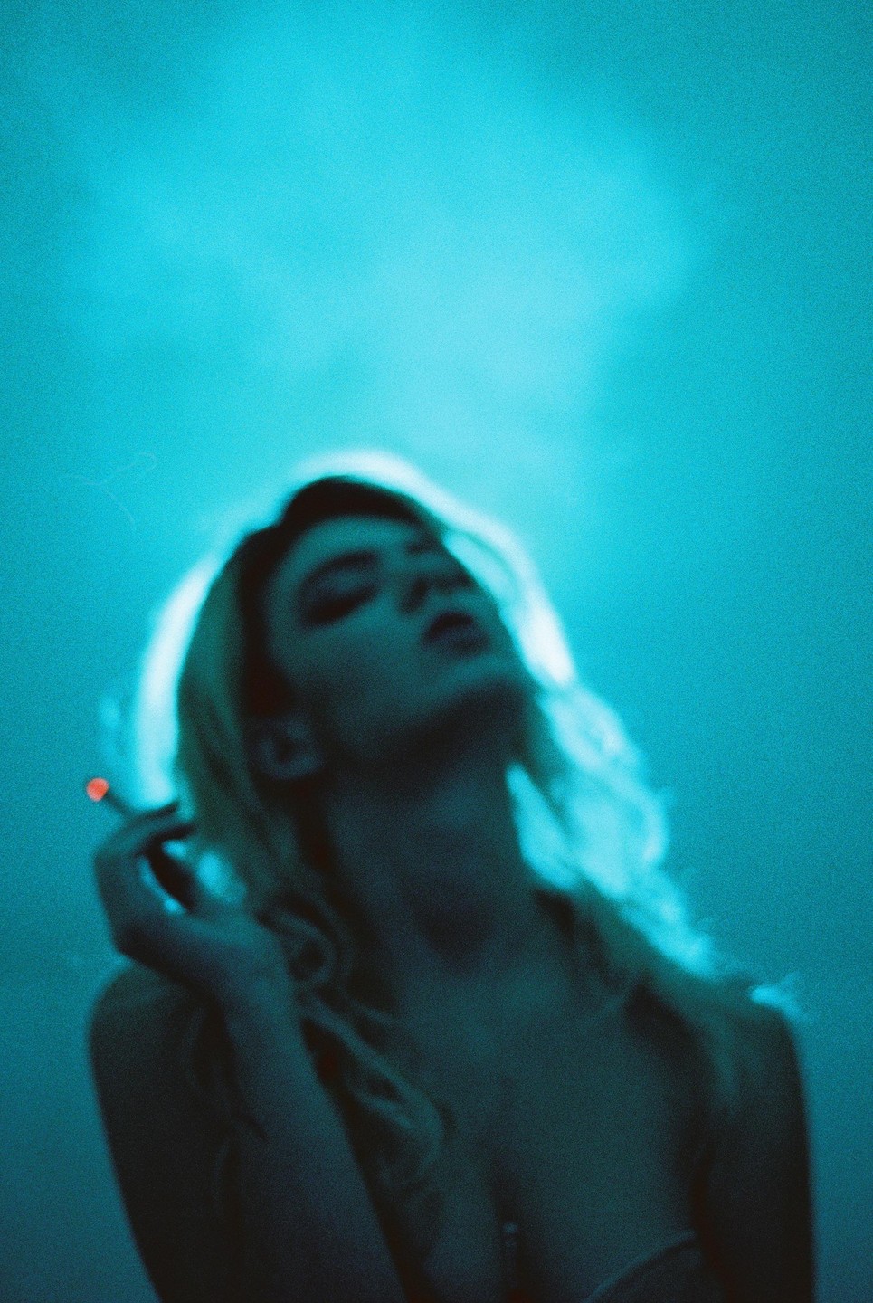 A woman with long hair holds a lit cigarette in a blue-lit room.