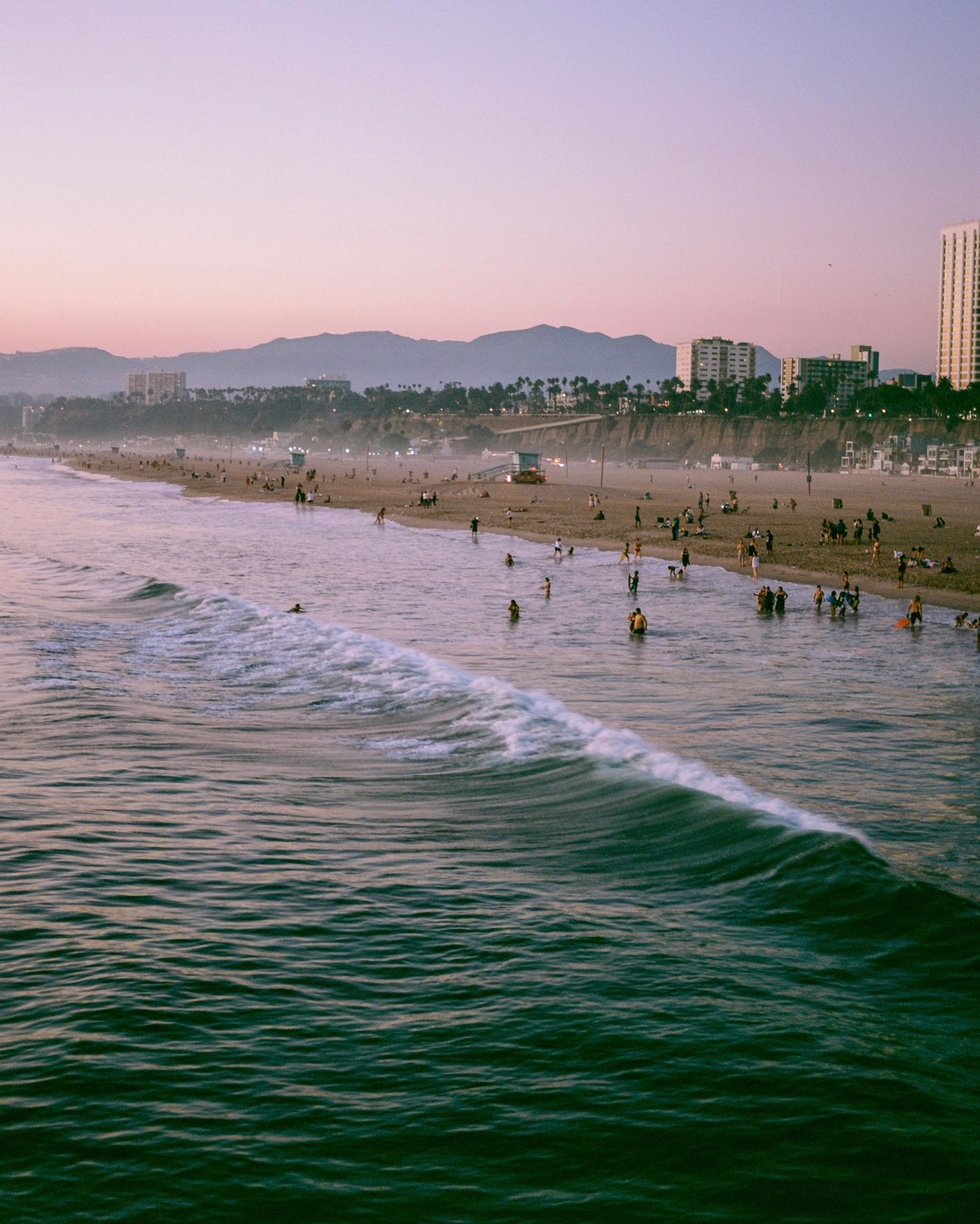 A wide shot shows a beach with many people, a city, and mountains in the distance under a pink sky.