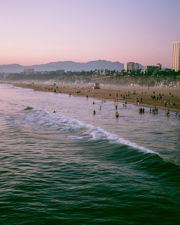 A wide shot shows a beach with many people, a city, and mountains in the distance under a pink sky.