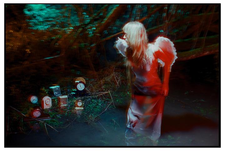 A woman with wings stands in water surrounded by clocks in a dark forest.