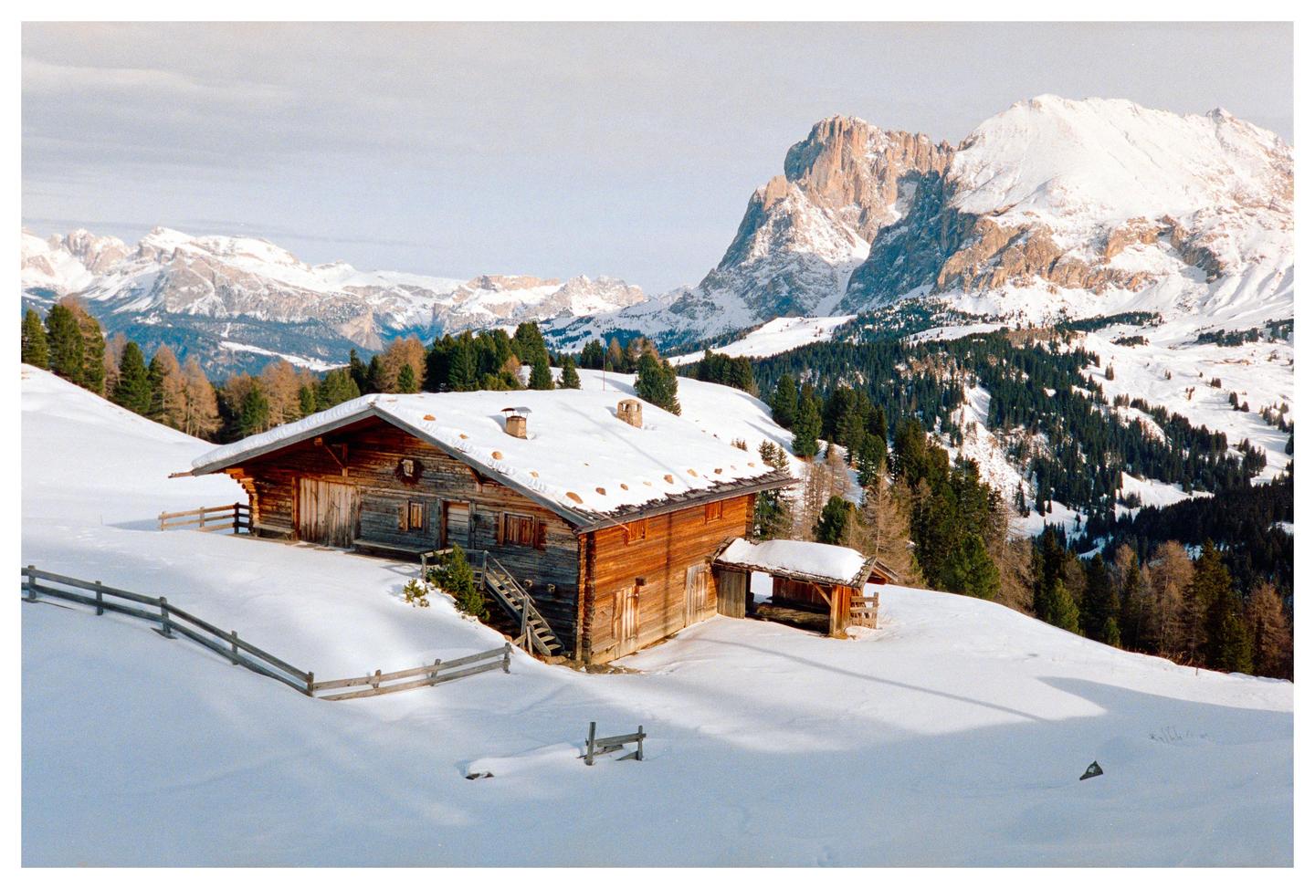 A wooden cabin with a snow-covered roof sits on a snowy hillside with mountains and trees in the background.