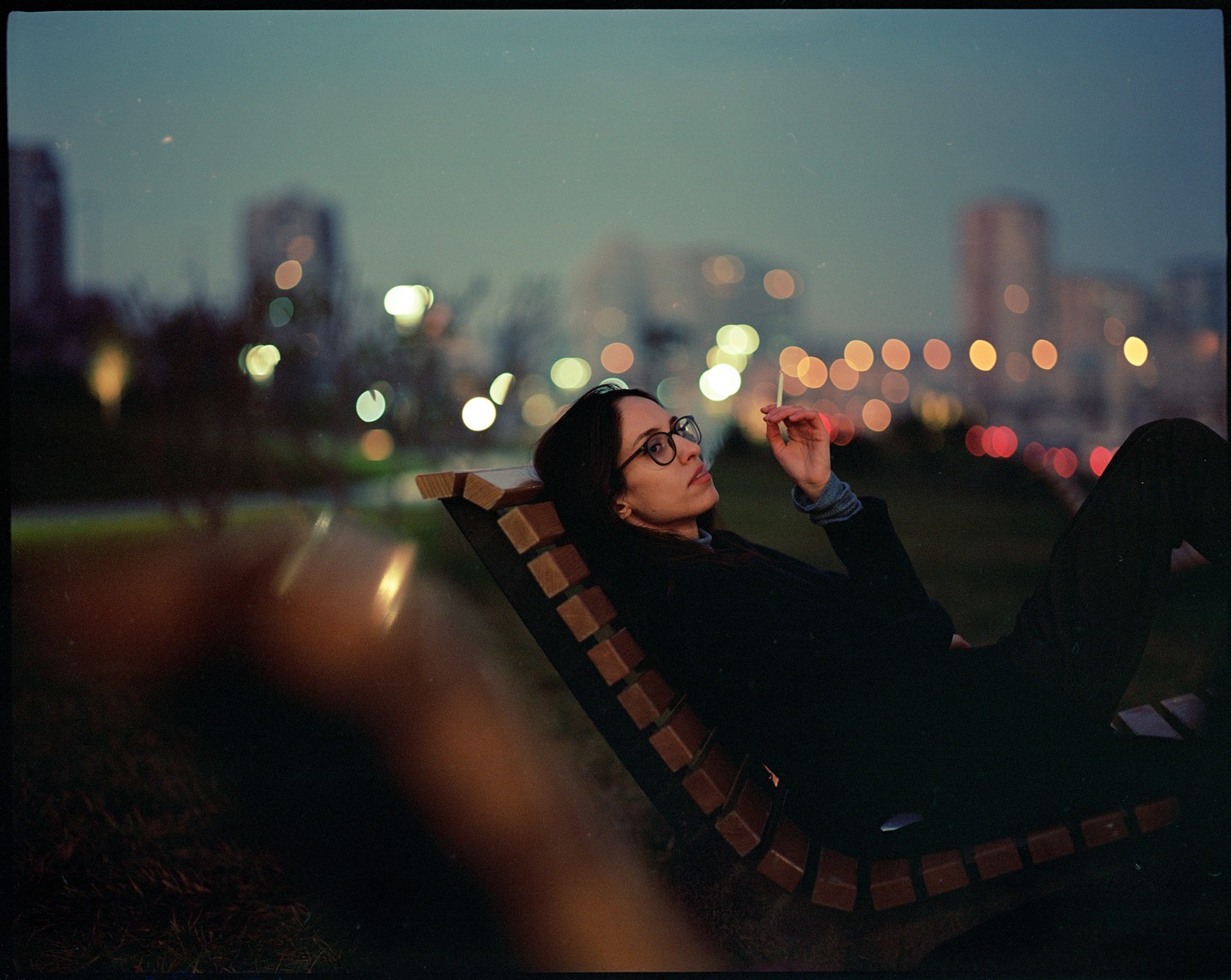A woman with glasses lies on a park bench and smokes a cigarette at night with city lights in the background.