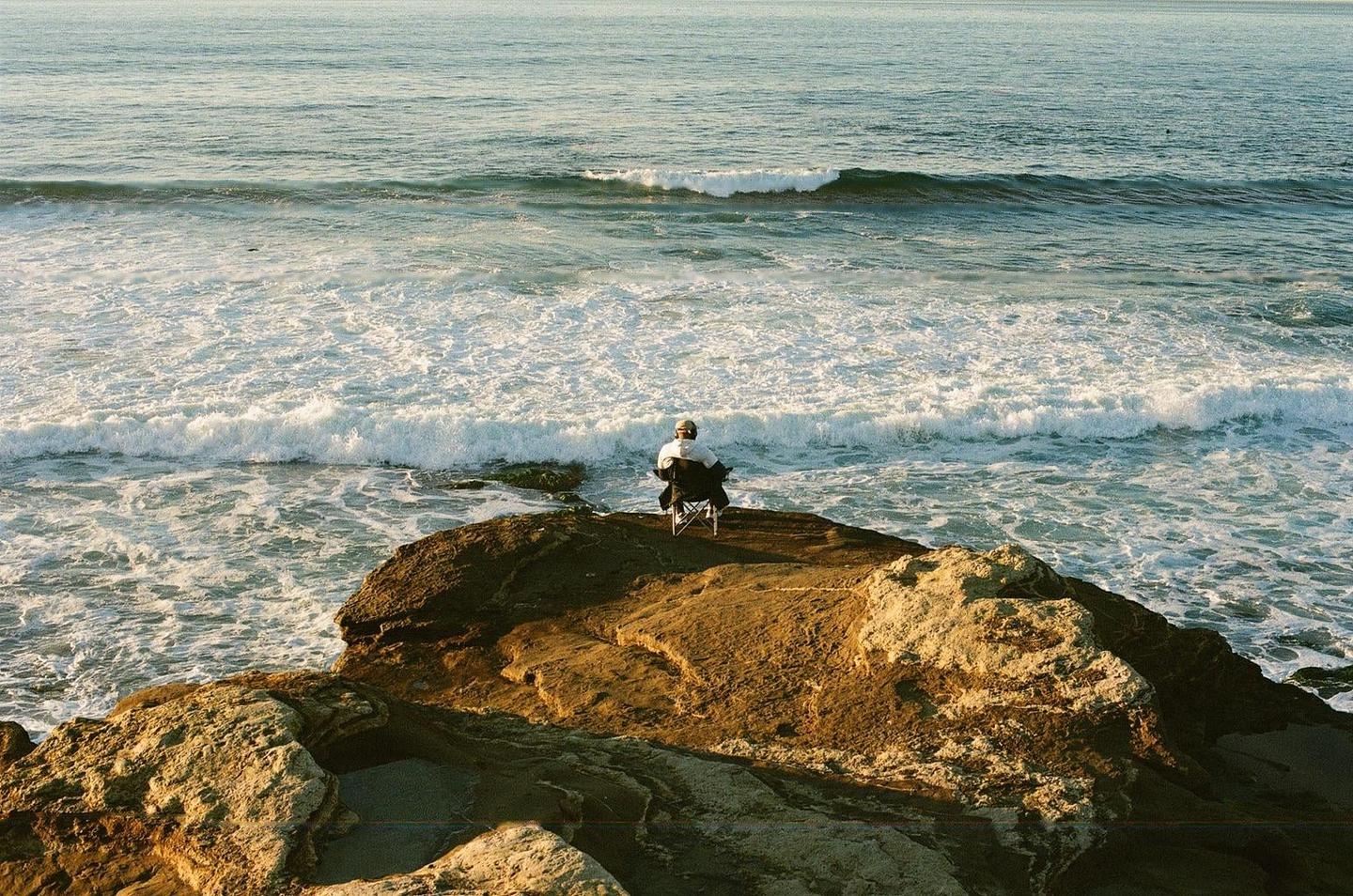 A person sits on a folding chair on a rocky cliff overlooking the ocean waves.