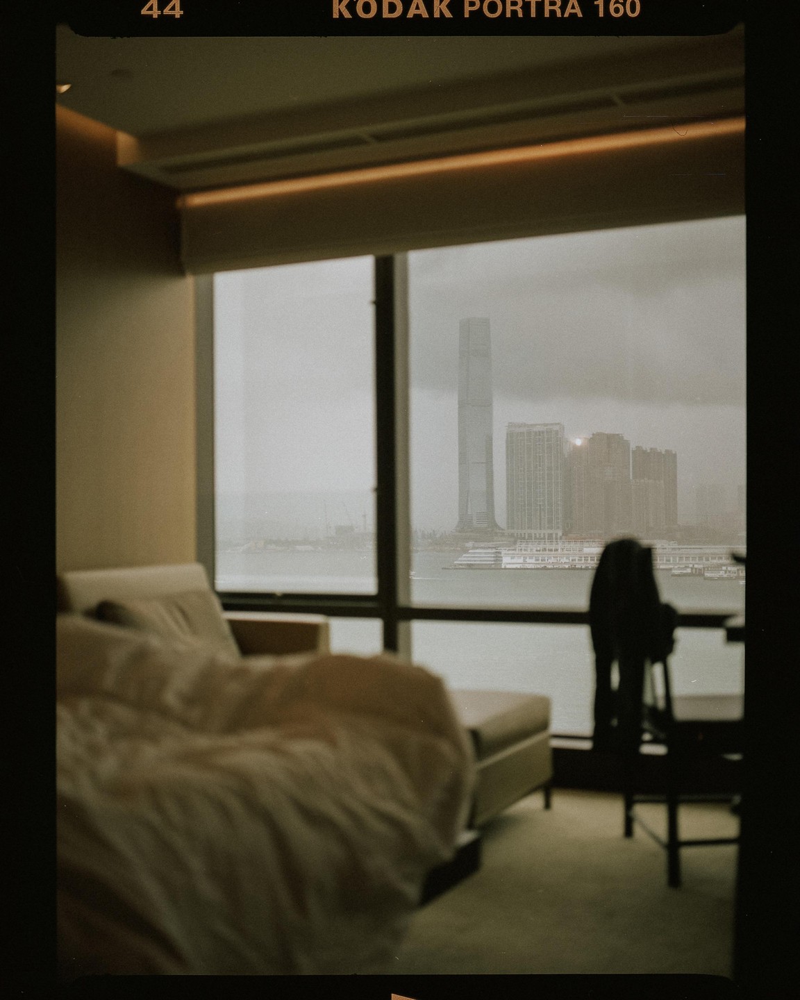 A bed and chaise lounge are in a room with a large window overlooking a body of water and a city skyline.