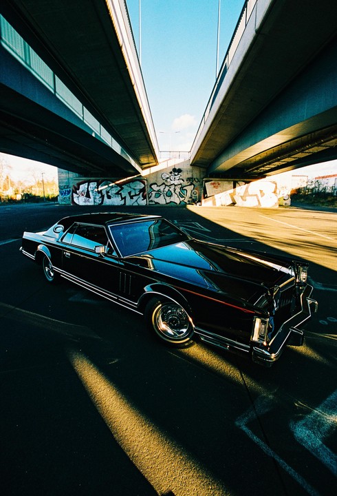 A black car is parked under a bridge with graffiti on the walls.