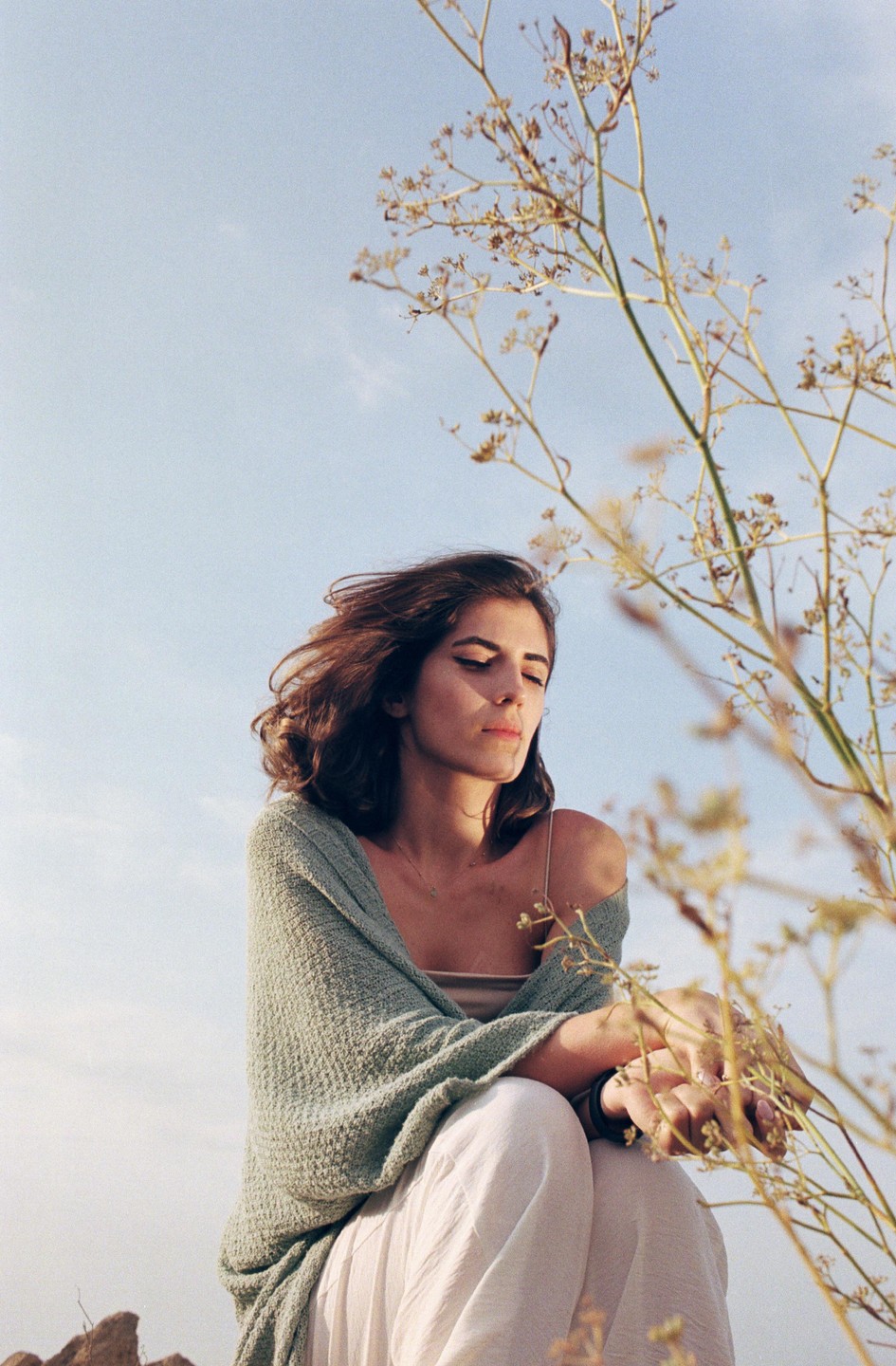 A woman with closed eyes is sitting outdoors with dry plants in the foreground and a clear sky in the background.
