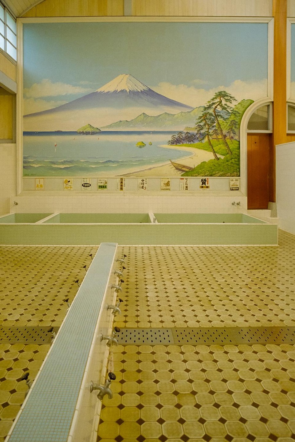 A large mural of Mount Fuji overlooking a beach and ocean is painted above several empty tiled bathing pools and a row of spigots in a public bathhouse.
