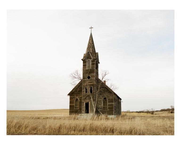 An old wooden church with a tall steeple stands in a field of dry grass under a pale sky.