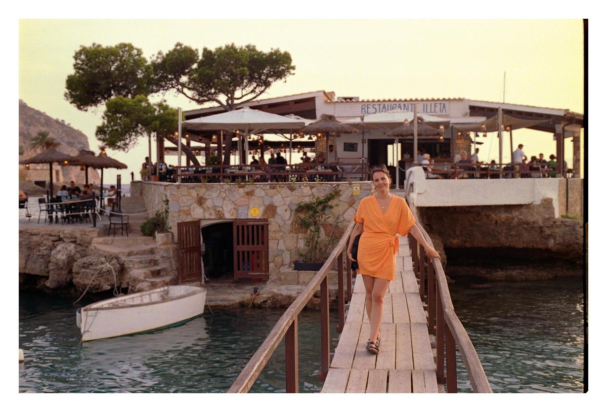 A woman in an orange dress walks on a wooden pier towards a restaurant on the water at dusk.