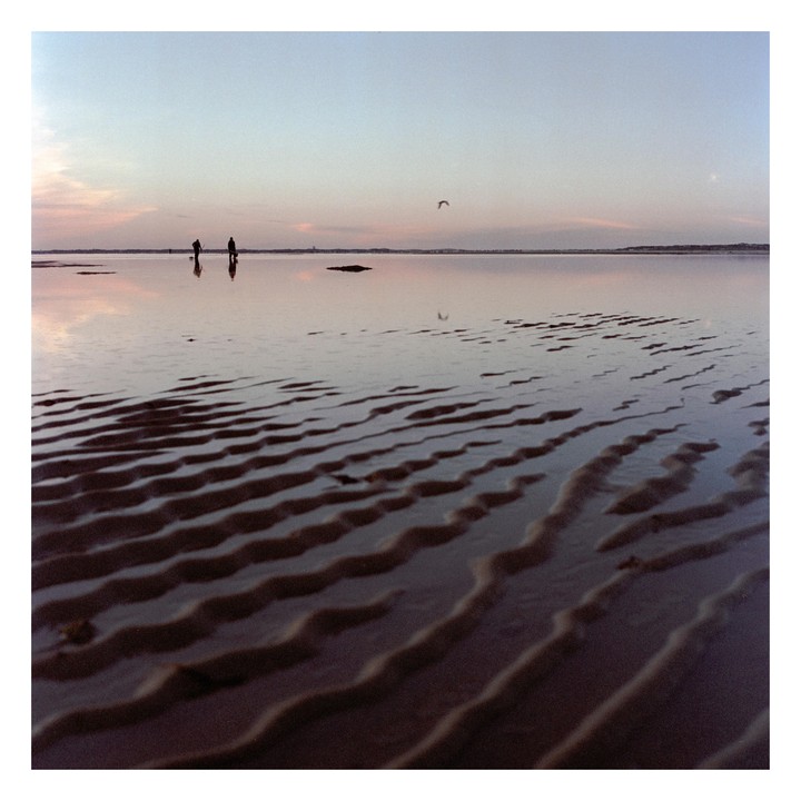 Two people walk on a sandy beach with water reflecting the sky at sunset.