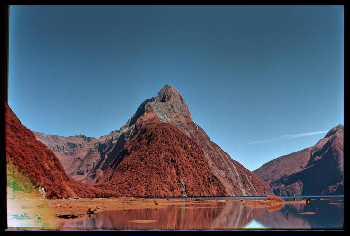 A large mountain with red foliage rises from a body of water, reflecting in the still surface under a clear sky.