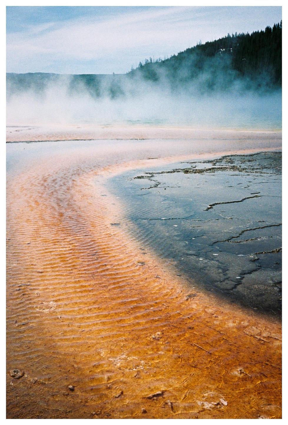A body of water with orange and dark blue sections is shown with steam rising in the background and a forest on a hill.
