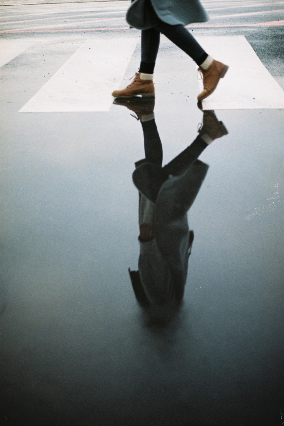 A person wearing boots walks across a wet crosswalk, with their reflection visible in the water.