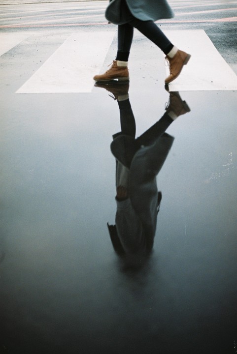 A person wearing boots walks across a wet crosswalk, with their reflection visible in the water.