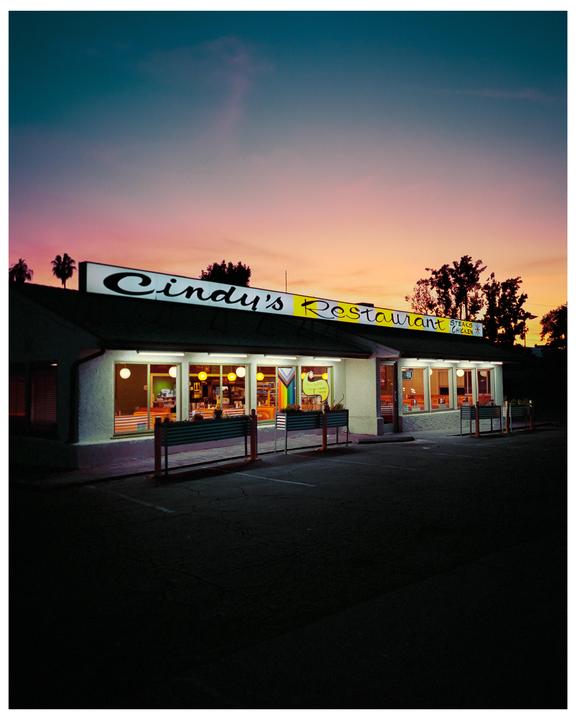 A restaurant with a brightly lit sign and interior sits under a colorful sunset sky.
