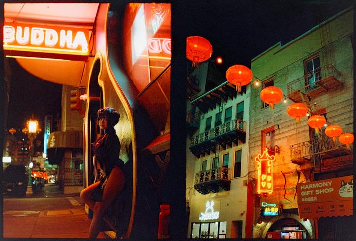 A woman in a dark outfit poses next to a neon sign that says "Buddha" while red lanterns hang above a building with balconies.