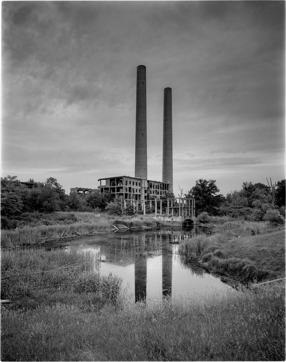 Two tall smokestacks rise above a derelict building, reflected in the still water of a pond surrounded by tall grass and trees.