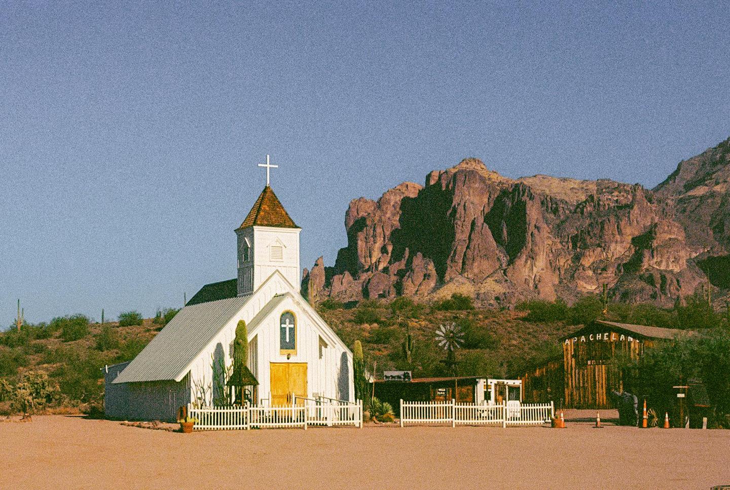 A white church with a steeple stands in a desert landscape with mountains in the background.