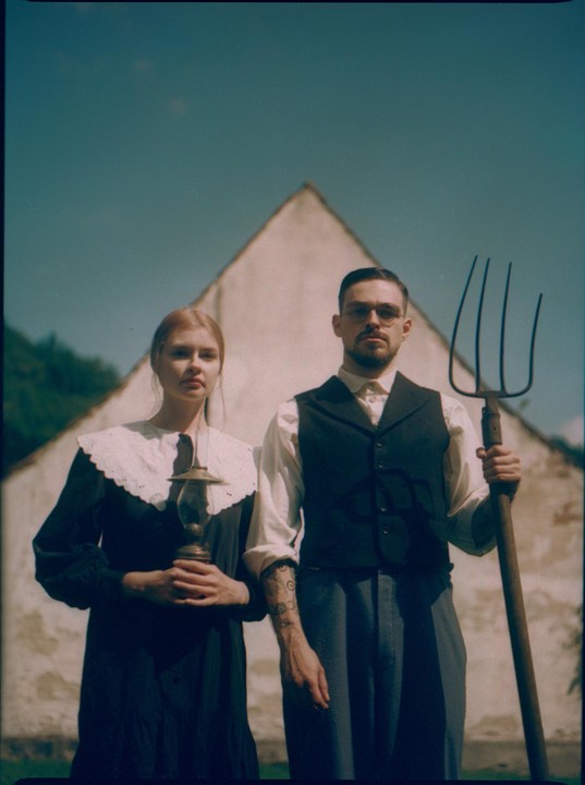 A woman holding a lantern and a man holding a pitchfork stand in front of a white building.
