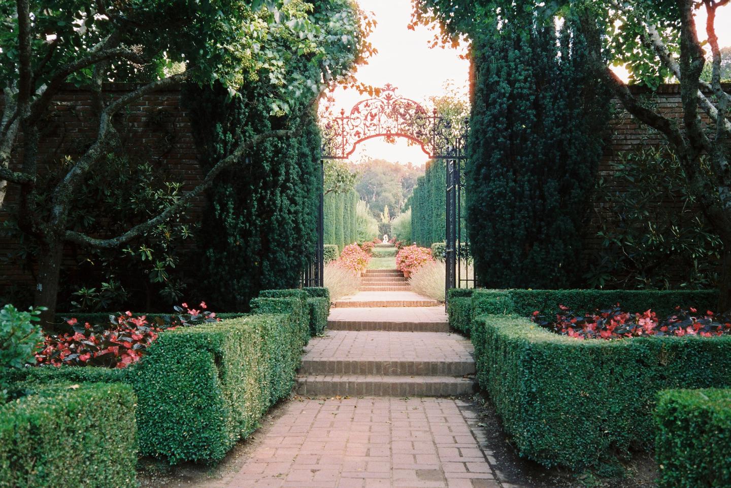A brick path leads through a formal garden with hedges, trees, and a decorative iron gate.