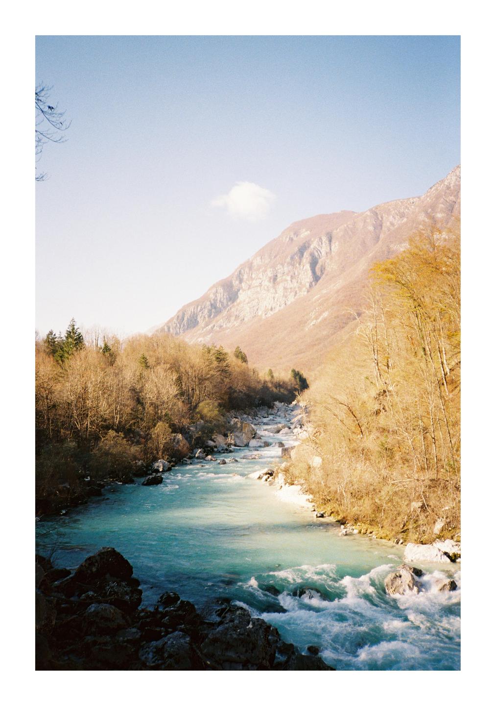 A turquoise river flows through a valley with mountains and bare trees under a clear sky.