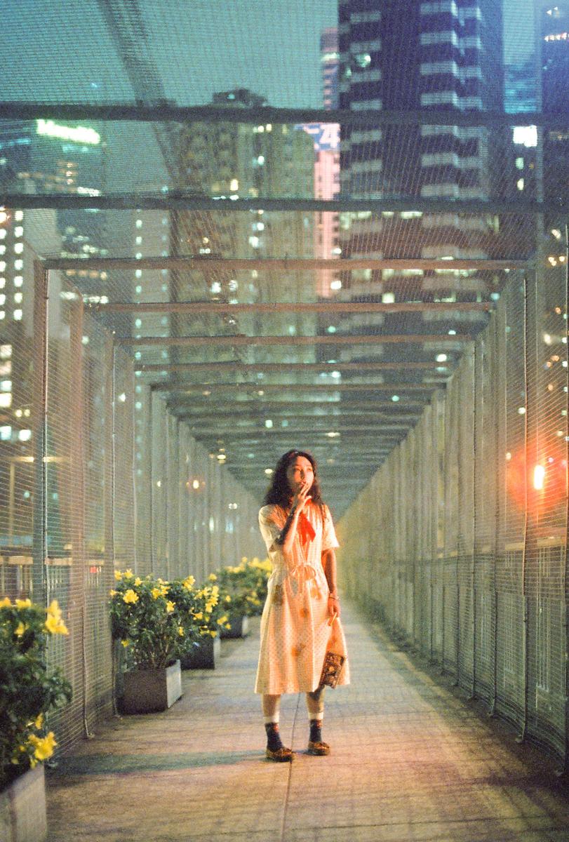 A woman stands in a covered walkway at night, surrounded by city lights and potted plants.