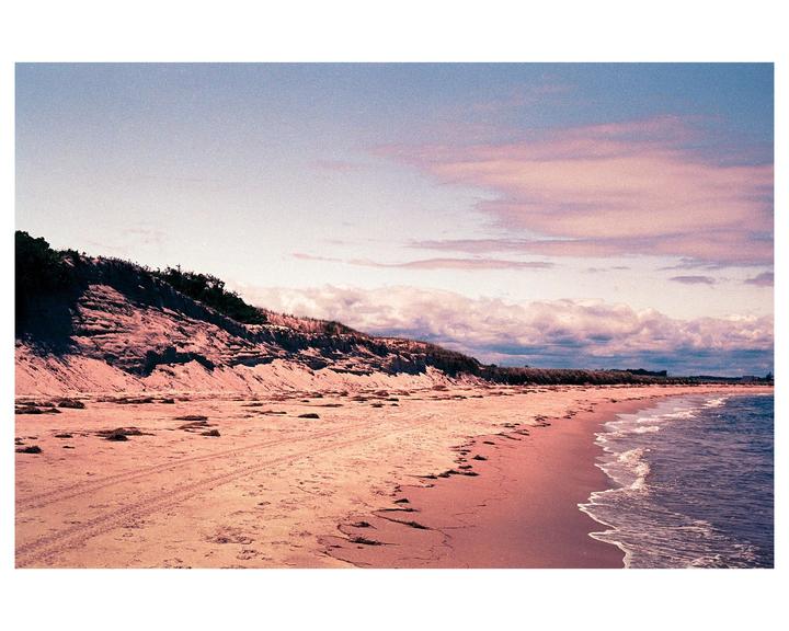 A sandy beach with a steep dune on the left and gentle waves on the right is visible under a cloudy sky.
