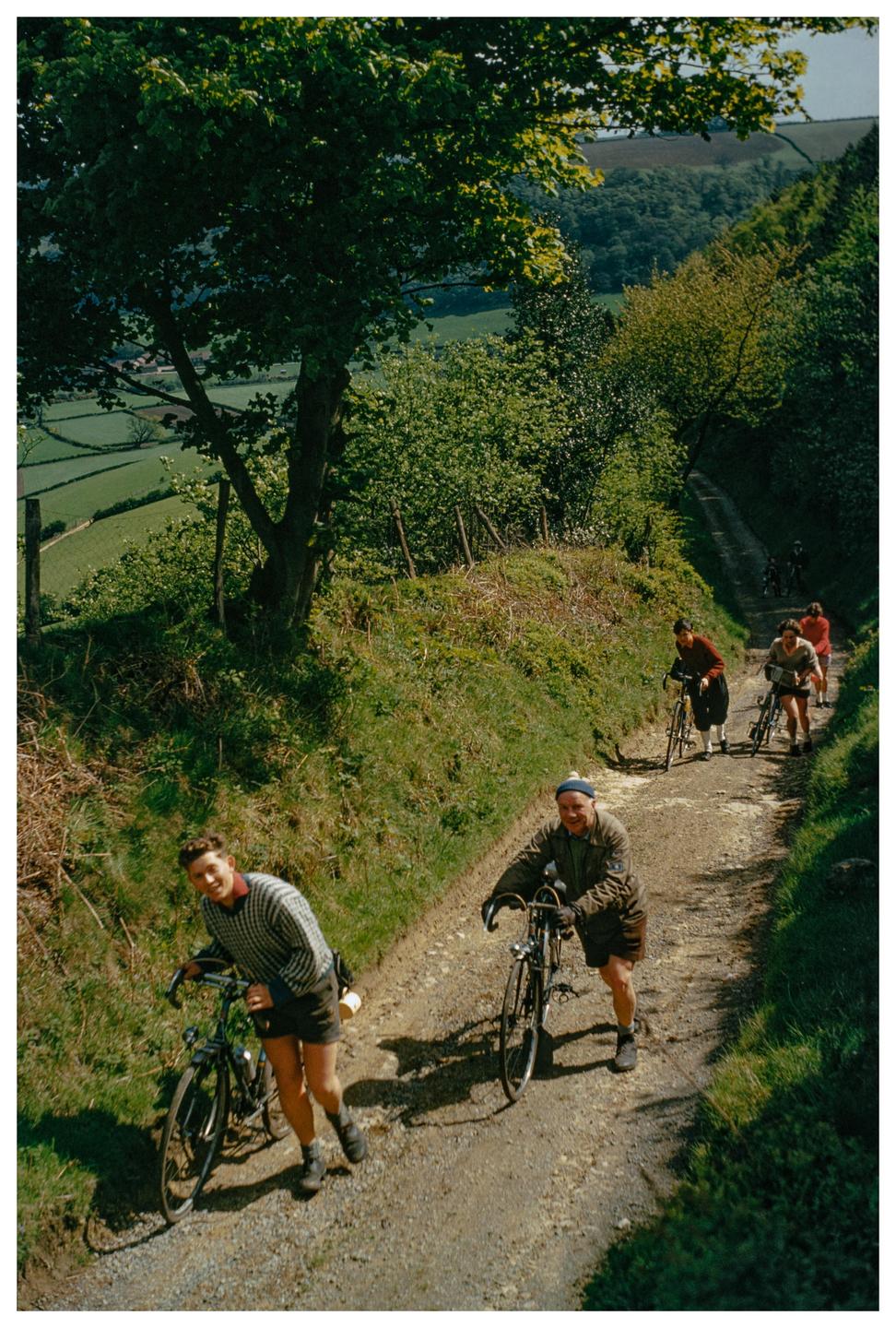 A group of cyclists push their bicycles up a steep, winding dirt path through a lush green landscape.