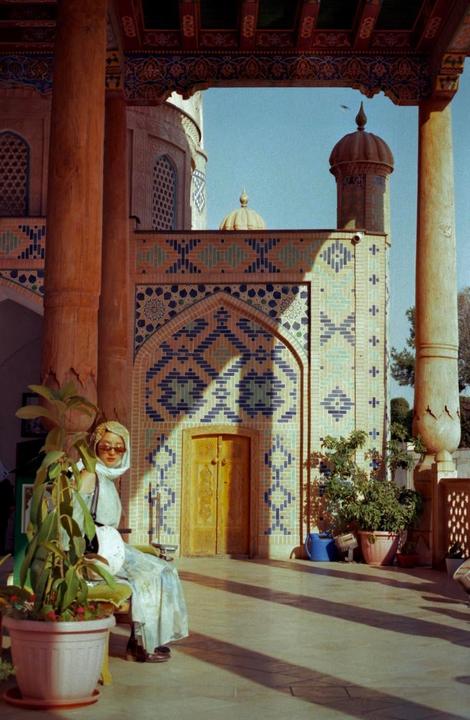 A woman in sunglasses and a headscarf sits on a bench next to a potted plant, with a building featuring patterned tiles and a wooden door in the background.