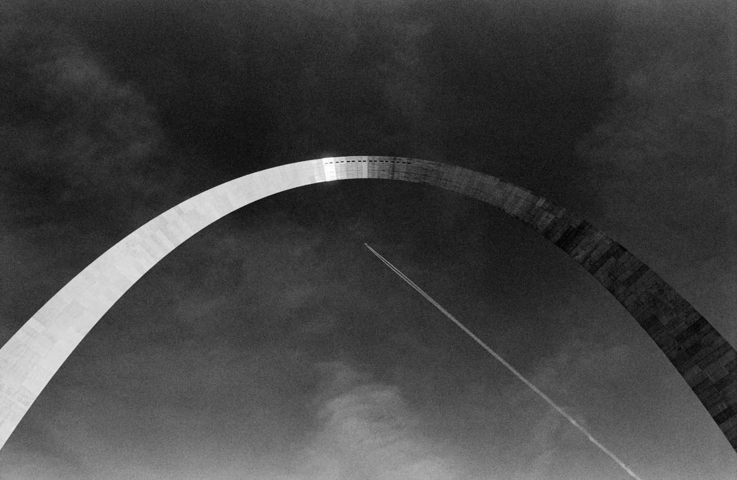 A high-contrast black and white image shows the Gateway Arch with an airplane contrail crossing the sky beneath it.