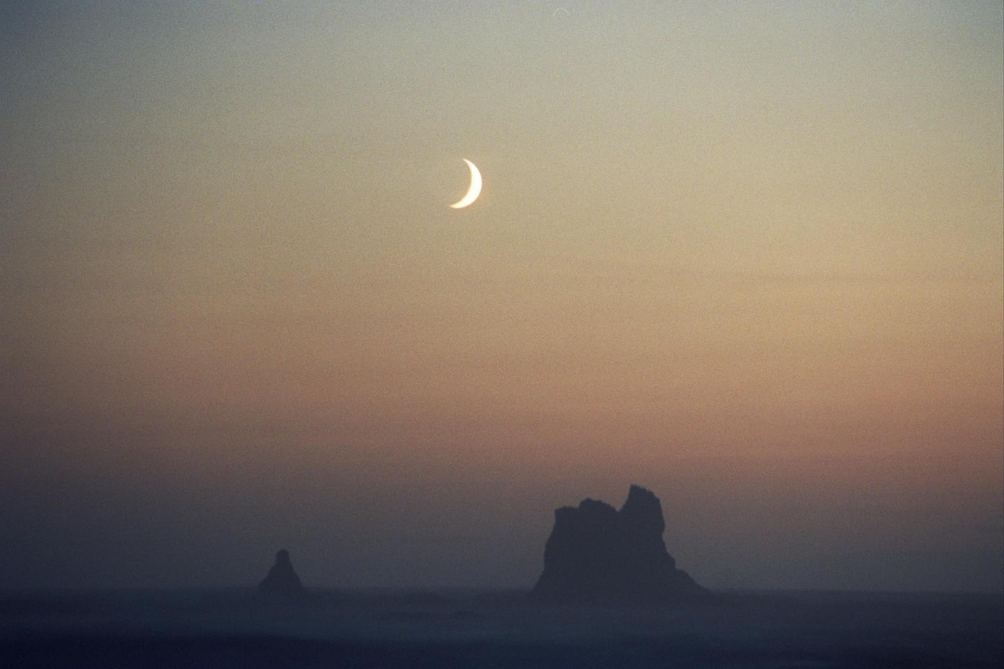 A crescent moon hangs in the sky above two dark rock formations on the Olympic Coast.