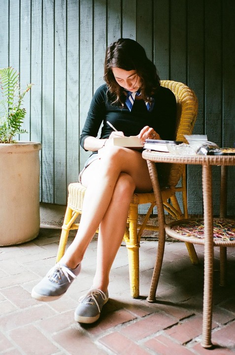 A woman sits in a wicker chair, writing in a book on a brick patio.