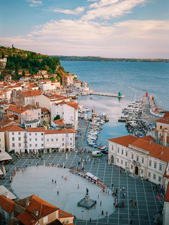 A coastal town with red-roofed buildings, a harbor filled with boats, and a bustling town square.