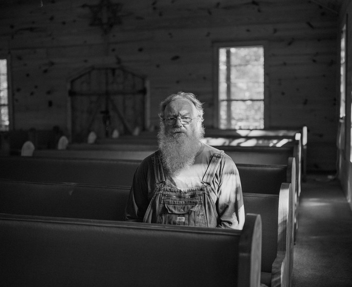 A man with a long beard and glasses sits in a church pew.