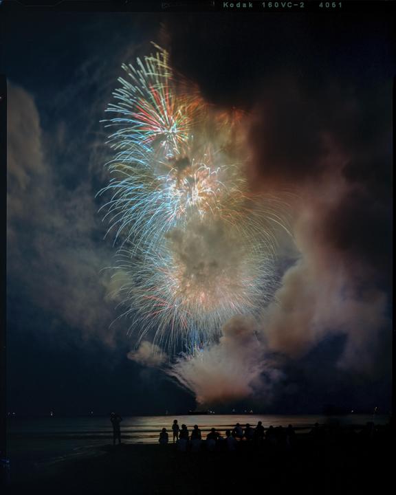 People watch colorful fireworks explode over a body of water at night.