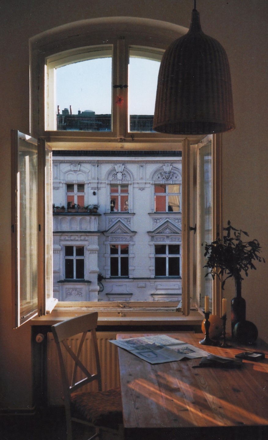 Sunlight streams through an open window, illuminating a wooden table with a newspaper and a chair in a room overlooking a building.