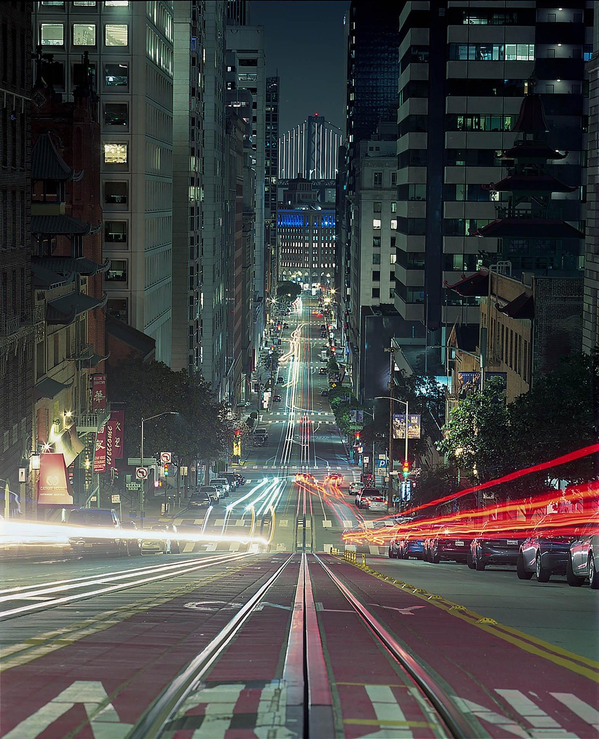 A long exposure shot of a city street at night shows light trails from vehicles and buildings lining the road.