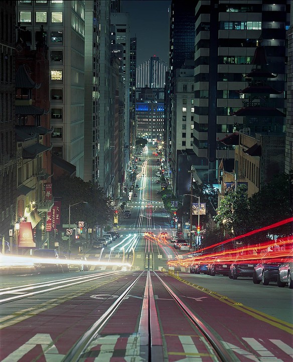 A long exposure shot of a city street at night shows light trails from vehicles and buildings lining the road.