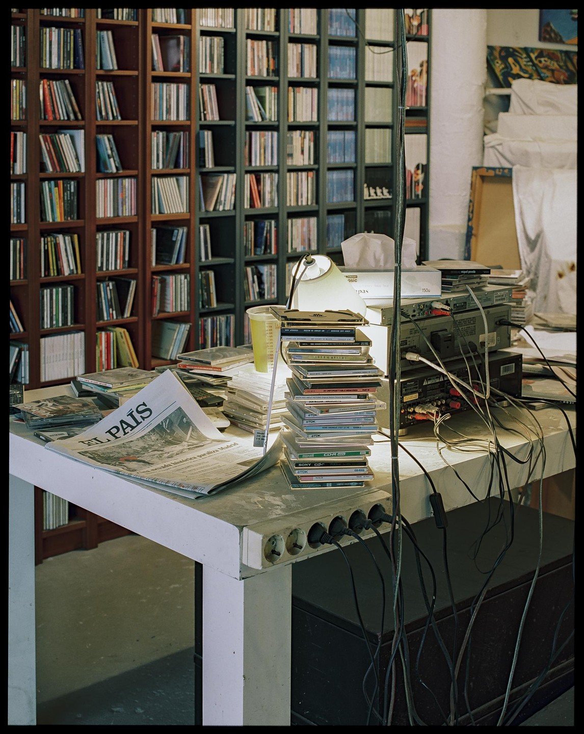 A newspaper, CDs, and electronics are on a table in front of bookshelves filled with CDs.