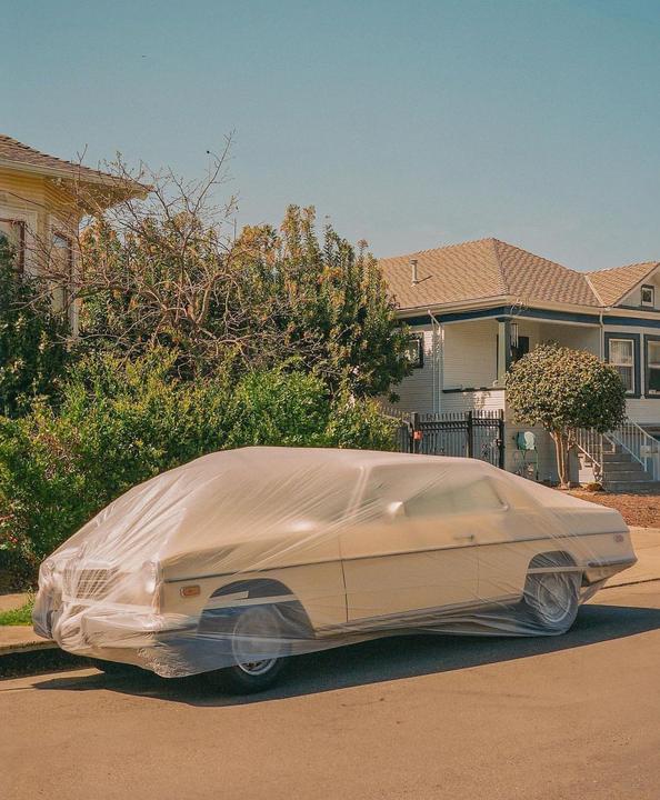 A car covered in plastic is parked on a residential street next to a sidewalk and houses.